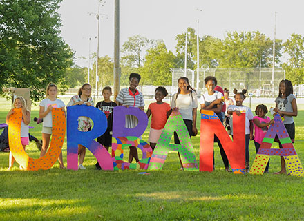 A group of children standing behind a set of letters that spell Urbana